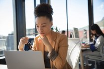 Eine Geschäftsfrau mit gemischter Rasse, die in einem modernen Büro arbeitet, am Schreibtisch sitzt und einen Laptop benutzt, während ihr Kollege im Hintergrund arbeitet. — Stockfoto