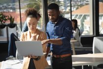 Eine Geschäftsfrau mit gemischter Rasse und ein afroamerikanischer Geschäftsmann arbeiten in einem modernen Büro, benutzen einen Laptop und unterhalten sich, während ihre Kollegen im Hintergrund arbeiten. — Stockfoto