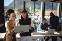 Смешанная раса и азиатские деловые женщины, работающие в современном офисе, используя ноутбук и разговаривая, со своими коллегами, работающими на заднем плане — стоковое фото