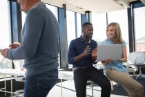 Uma mulher de negócios caucasiana e um empresário afro-americano trabalhando em um escritório moderno, usando um computador portátil e conversando, com seu colega trabalhando em primeiro plano — Fotografia de Stock