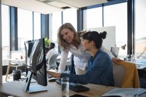 Eine kaukasische und eine asiatische Geschäftsfrau arbeiten in einem modernen Büro, benutzen einen Laptop und unterhalten sich, während ihre Kollegen im Hintergrund arbeiten — Stockfoto