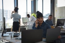 Азиатская деловая женщина, работающая в современном офисе, сидящая за столом, используя ноутбук и разговаривая на смартфоне, со своими коллегами, работающими на заднем плане — стоковое фото