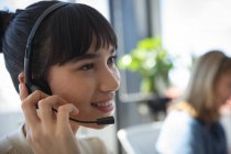Großaufnahme einer asiatischen Geschäftsfrau, die in einem modernen Büro arbeitet, am Schreibtisch sitzt, Headset trägt und spricht, während ihr Kollege im Hintergrund arbeitet — Stockfoto