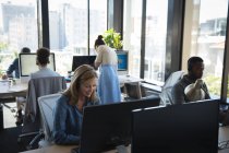 Багатоетнічна група чоловічих і жіночих колег, що працюють в сучасному офісі, сидять за столами, використовують комп'ютери, носять навушники і розмовляють — стокове фото