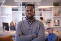 Портрет афроамериканського бізнесмена, який працює в сучасному офісі, дивиться на камеру зі схрещеними руками, з колегами на задньому плані. — стокове фото