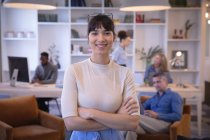 Porträt einer glücklichen asiatischen Geschäftsfrau, die in einem modernen Büro arbeitet, in die Kamera blickt und lächelt, während ihre Kollegen im Hintergrund arbeiten — Stockfoto