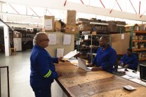 Un supervisor masculino de raza mixta y un trabajador afroamericano en un almacén en una fábrica que fabrica sillas de ruedas, de pie y hablando en un banco de trabajo - foto de stock
