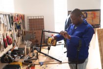 Un trabajador afroamericano en un taller en una fábrica que fabrica sillas de ruedas, parado en un banco de trabajo usando una herramienta de mano y ensamblando partes de un producto - foto de stock