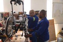 Trabajadores afroamericanos y de raza mixta en un taller en una fábrica que fabrica sillas de ruedas, de pie en un banco de trabajo y ensamblando partes de un producto - foto de stock