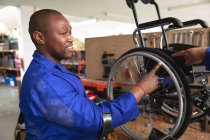 Un trabajador afroamericano en un taller en una fábrica que fabrica sillas de ruedas, parado en un banco de trabajo y ensamblando partes de un producto - foto de stock