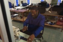 Um trabalhador afro-americano em uma oficina em uma fábrica de cadeiras de rodas, sentado em uma bancada, usando uma máquina de costura — Fotografia de Stock