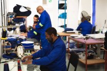 Um trabalhador afro-americano em uma oficina em uma fábrica de cadeiras de rodas, sentado em uma bancada de trabalho, usando uma máquina de costura, com outros trabalhadores trabalhando em segundo plano — Fotografia de Stock