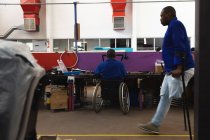 Disabled African American male worker in a workshop at a factory making wheelchairs, sitting at a workbench assembling parts of a product, sitting in wheelchair, a colleague walking past on crutches — Stock Photo
