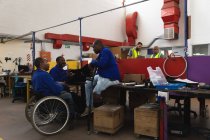 Group of disabled African American male workers in a workshop at a factory making wheelchairs, sitting at a workbench assembling parts of a product, one sitting in a wheelchair, one using crutches — Stock Photo