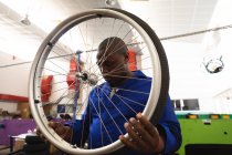 Um trabalhador afro-americano na oficina em uma fábrica de cadeiras de rodas, de pé e inspecionando uma roda, vestindo uma roupa de trabalho — Fotografia de Stock