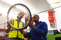 An African American male worker and a Caucasian male supervisor in the workshop at a factory making wheelchairs, standing and inspecting a wheel together, wearing a workwear — Stock Photo