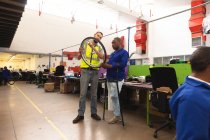 Um trabalhador afro-americano do sexo masculino e um supervisor caucasiano do sexo masculino na oficina em uma fábrica de cadeiras de rodas, de pé e inspecionando uma roda juntos, vestindo uma roupa de trabalho — Fotografia de Stock