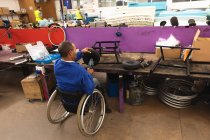 Trabajador afroamericano discapacitado en un taller en una fábrica que fabrica sillas de ruedas, sentado en un banco de trabajo ensamblando partes de un producto, sentado en silla de ruedas - foto de stock