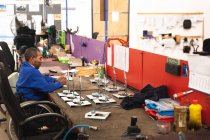 Афроамериканський робітник - інвалід на фабриці, який виготовляє інвалідні візки, сидить на робочому місці і збирає частини продукту, сидячи в інвалідному візку. — стокове фото