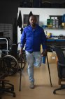 Retrato de um trabalhador afro-americano deficiente com uma perna de pé usando muletas vestindo roupas de trabalho, em um armazém de armazenamento em uma fábrica fazendo cadeiras de rodas, olhando para a câmera — Fotografia de Stock