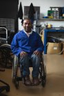 Porträt eines behinderten Arbeiters mit gemischter Rasse, der Arbeitskleidung trägt, in einer Lagerhalle einer Fabrik, die Rollstühle herstellt, in die Kamera blickt und im Rollstuhl sitzt — Stockfoto