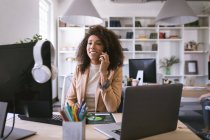 Una mujer de negocios de raza mixta que trabaja en una oficina moderna, sentada en un escritorio y usando una computadora, hablando en un teléfono inteligente, con sus colegas de negocios trabajando en segundo plano - foto de stock
