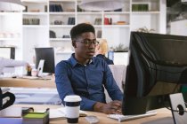 Un homme d'affaires afro-américain travaillant dans un bureau moderne, assis à un bureau et utilisant un ordinateur, avec ses collègues d'affaires travaillant en arrière-plan — Photo de stock