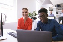 Eine lächelnde kaukasische Geschäftsfrau und ein afroamerikanischer Geschäftsmann arbeiten in einem modernen Büro, benutzen einen Laptop und unterhalten sich, während ihre Geschäftskollegen im Hintergrund arbeiten. — Stockfoto