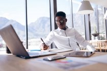 Um empresário afro-americano trabalhando em um escritório moderno, sentado em uma mesa perto de uma janela em um dia ensolarado usando um computador portátil, segurando uma caneta e tomando notas — Fotografia de Stock
