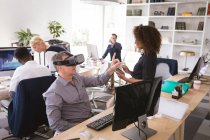 Um empresário caucasiano trabalhando em um escritório moderno, vestindo um fone de ouvido VR e tocando tela interativa virtual, com seus colegas de negócios trabalhando em mesas em segundo plano — Fotografia de Stock