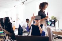 Кавказская деловая женщина, работающая в современном офисе, в VR гарнитуре, трогательная виртуальный интерактивный экран, со своими деловыми коллегами, работающими на заднем плане — стоковое фото