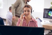 Кавказька бізнесменка, яка працює в сучасному офісі, сидить за столом, користується ноутбуком, носить навушники і розмовляє зі своїми колегами на задньому плані. — стокове фото