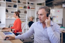 Un homme d'affaires caucasien travaillant dans un bureau moderne, assis à un bureau, portant un casque et parlant au téléphone, avec ses collègues d'affaires travaillant en arrière-plan — Photo de stock