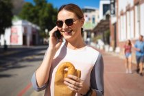 Una mujer de negocios caucásica en movimiento en un día soleado, de pie en una calle de la ciudad, sosteniendo un bagel y hablando en su teléfono inteligente, usando gafas de sol - foto de stock