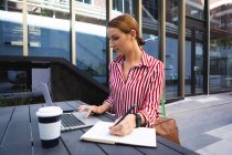 Una mujer de negocios caucásica en movimiento en un día soleado, sentada en una mesa, usando su computadora portátil y tomando notas, con un café para llevar en una mesa - foto de stock