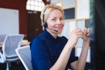 Porträt einer kaukasischen Geschäftsfrau mit kurzen blonden Haaren, die in einem modernen Büro arbeitet, am Schreibtisch sitzt, Headset trägt und in die Kamera blickt — Stockfoto