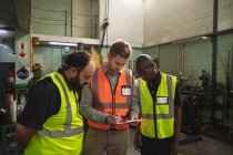 Two Caucasian and an African American male factory workers wearing a high vis vest talking and holding clipboard. Workers in industry at a factory making hydraulic equipment. — Stock Photo