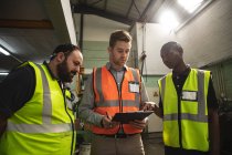 Two Caucasian and an African American male factory workers wearing a high vis vest talking and holding clipboard. Workers in industry at a factory making hydraulic equipment. — Stock Photo