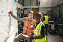 Caucasian and African American male factory workers wearing a high vis vest holding clipboard and writing on a board, talking. Workers in industry at a factory making hydraulic equipment. — Stock Photo