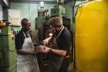 Afroamerikanische und kaukasische männliche Fabrikarbeiter tragen Schürze, reden und halten hydraulische Teile. Arbeiter in der Industrie in einer Fabrik für hydraulische Anlagen. — Stockfoto