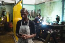 African American male factory worker wearing apron using smartphone with coworker welding in the background. Workers in industry at a factory making hydraulic equipment. — Stock Photo