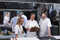 Caucasian female chef holding a file of papers, talking to Caucasian male and female chefs, standing by her with other chefs standing in the background. — Stock Photo