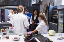 Mixed race waitress, writing on a notebook, talking to two Caucasian female chefs with other chefs standing in the background. Cookery class at a restaurant kitchen. — Stock Photo
