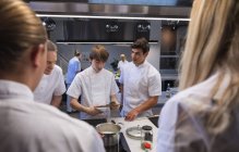 Three Caucasian male chefs standing by a table, looking at pans and pots, talking to each other, with Caucasian female chefs in the foreground. Cookery class at a restaurant kitchen. — Stock Photo