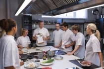 Grupo caucasiano de chefs masculinos e femininos, ouvindo um chef caucasiano sênior segurando um arquivo de papéis. Aula de culinária em uma cozinha de restaurante. — Fotografia de Stock