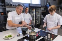 Grupo caucasiano de chefs masculinos e femininos, ouvindo um chef caucasiano sênior adicionando ingredientes a um pote. Aula de culinária em uma cozinha de restaurante. — Fotografia de Stock