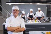 Retrato de un chef caucásico cruzando los brazos, mirando a la cámara y sonriendo, con otros chefs cocinando al fondo. Clase de cocina en una cocina de restaurante. - foto de stock