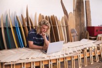 Creatore di tavole da surf caucasiche nel suo studio, che lavora a un progetto usando il suo portatile, con tavole da surf in un rack sullo sfondo. Tecnologia sportiva per le piccole imprese on line. — Foto stock