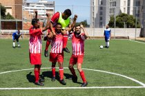 Zwei multiethnische Mannschaften von fünf männlichen Fußballern pro Seite tragen einen Mannschaftsstreifen bei einem Spiel auf einem Sportplatz in der Sonne und feiern den Sieg mit einem Spieler. — Stockfoto