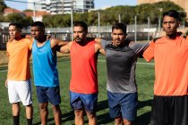 Multi grupo étnico de cinco hombres un lado jugadores de fútbol con entrenamiento de ropa deportiva en un campo de deportes en el sol, de pie en una fila abrazando antes de un juego. - foto de stock